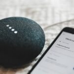 Charcoal smart speaker with glowing lights next to a smartphone, illustrating AI voice assistants